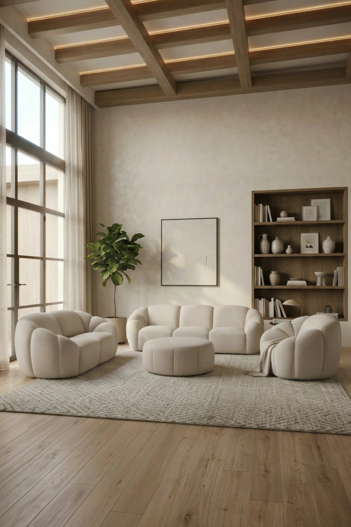 A modern living room featuring a cohesive set of cream-colored modular furniture, including a curved sofa, two matching armchairs, a round ottoman, and a textured area rug. The room has light wood floors, a high ceiling with exposed beams and recessed lighting, a large window on the left, and a wooden bookshelf on the right.