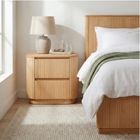 A light natural wood nightstand with two drawers and rounded edges, styled next to a bed with white linens and a green throw blanket in a bright bedroom.