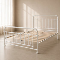 A white metal bed frame with wooden slats, shown in a softly lit beige room with curtains and sunlight streaming in.