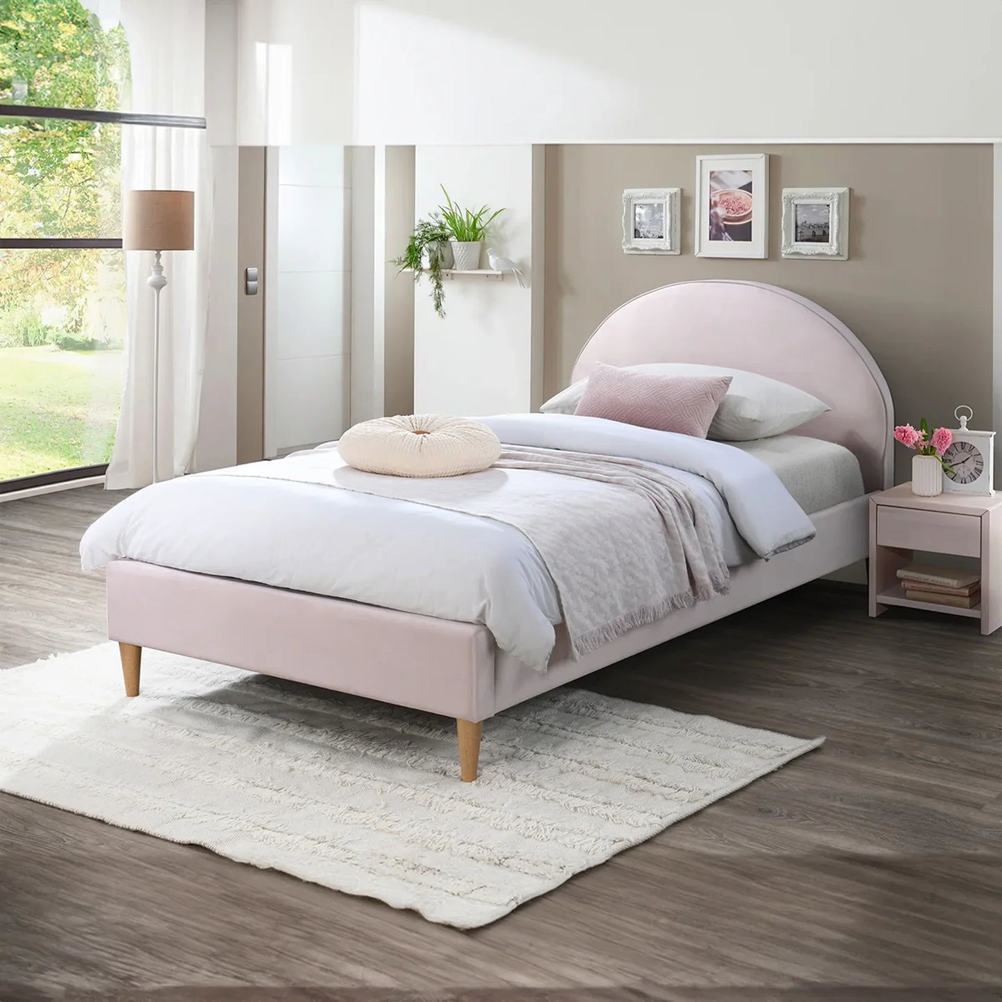 A light pink upholstered bed frame with a curved headboard and wooden legs, styled in a modern bedroom setting with white bedding, a small pink nightstand, and a white rug.