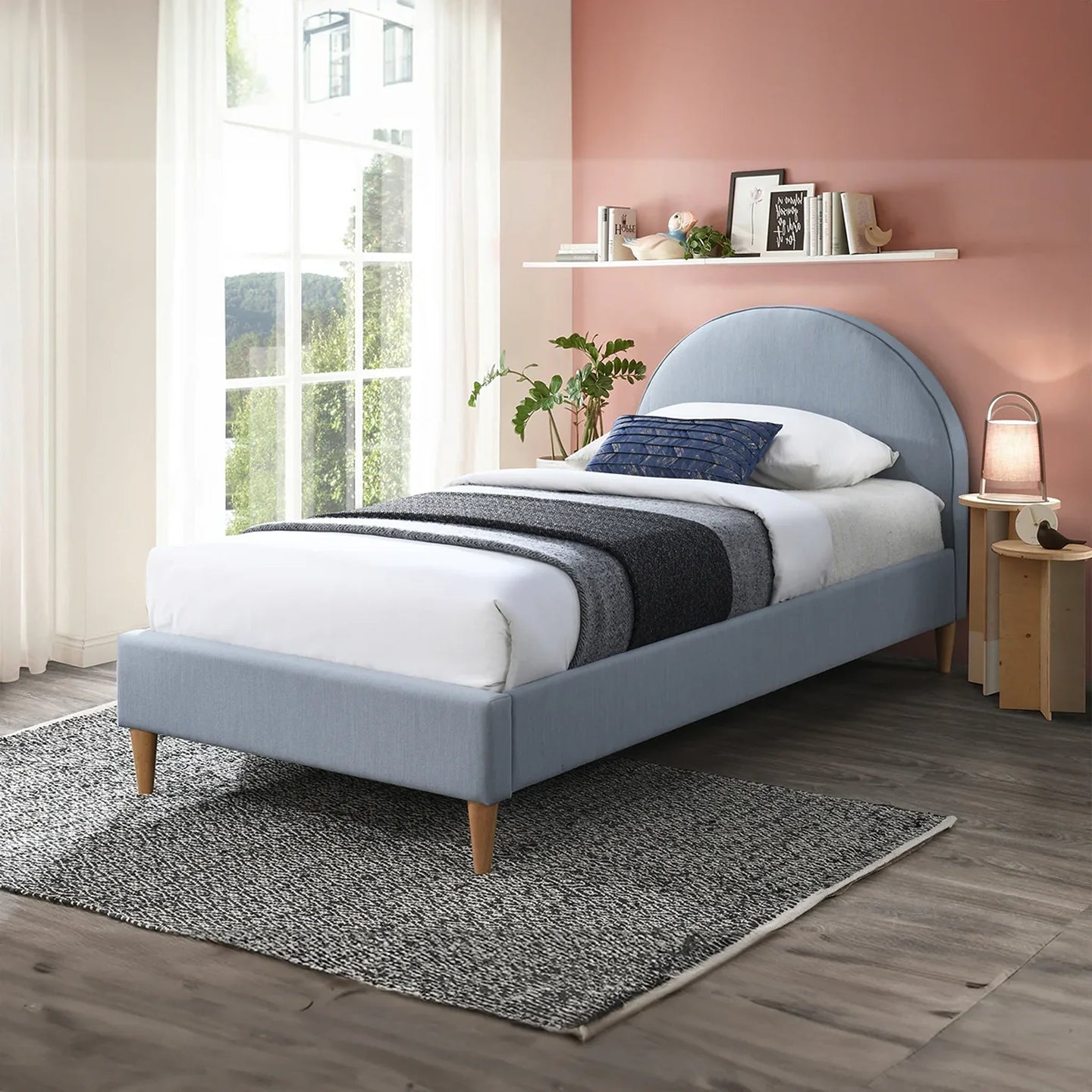 A light blue upholstered single bed with a curved headboard and wooden legs, styled in a modern bedroom with a pink wall, white curtains, a gray rug, and a bedside lamp.