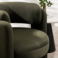 A close-up of a modern armchair upholstered in dark olive green textured fabric, shown in a styled interior setting with a side table and plant in the background.
