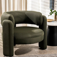 An olive green textured armchair with a curved, modern silhouette, set in a styled living room next to a black side table with books and a plant.