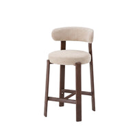 A modern bar stool with a round upholstered seat and curved backrest in a light beige fabric, supported by four dark brown wooden legs with a footrest.