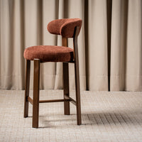 A modern bar stool with a rounded, upholstered backrest and seat cushion in a rust-orange textured fabric, supported by a dark brown wooden frame.