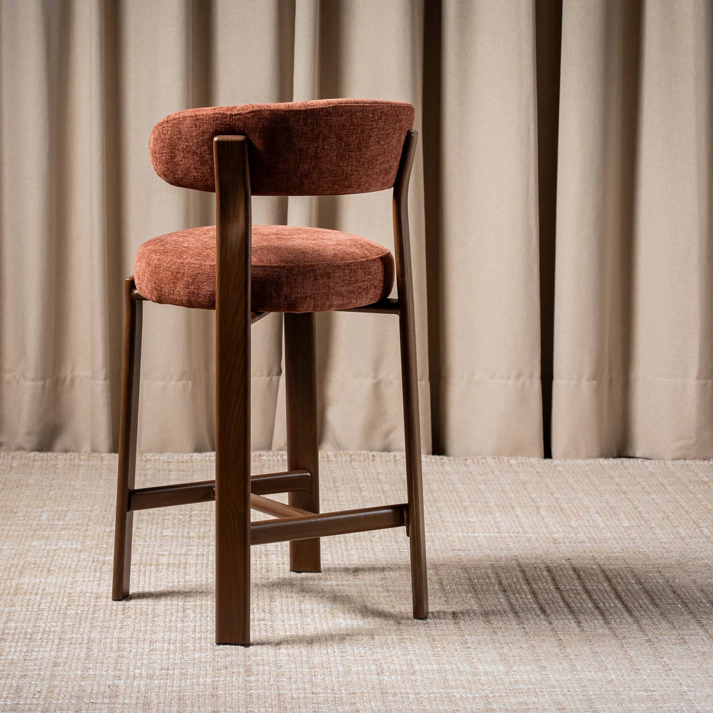 A modern bar stool with a rounded, cushioned backrest and seat upholstered in a textured rust-orange fabric, supported by a dark brown wooden frame