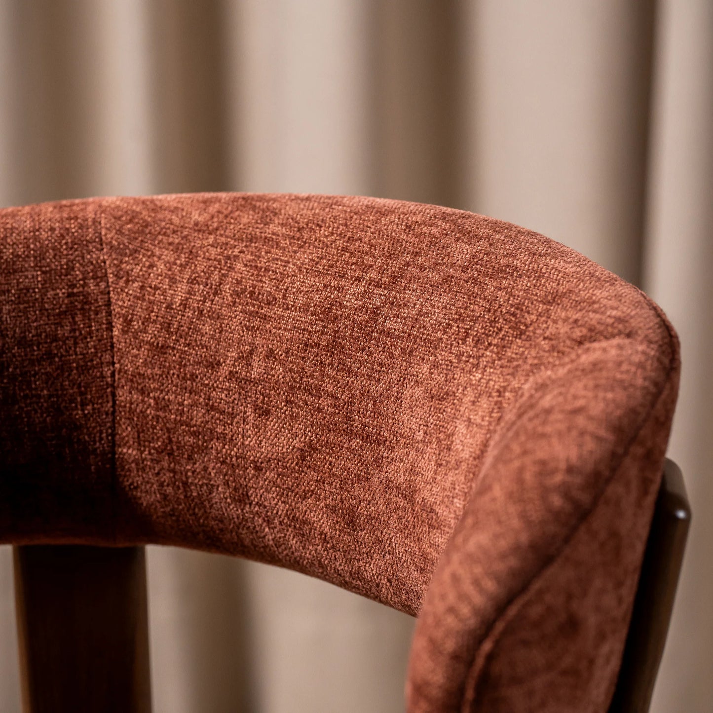 Close-up of a curved upholstered chair back in a warm rust-brown textured fabric, with a dark wooden leg