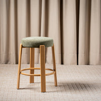 A modern bar stool with a round sage green upholstered cushion and light natural wood legs