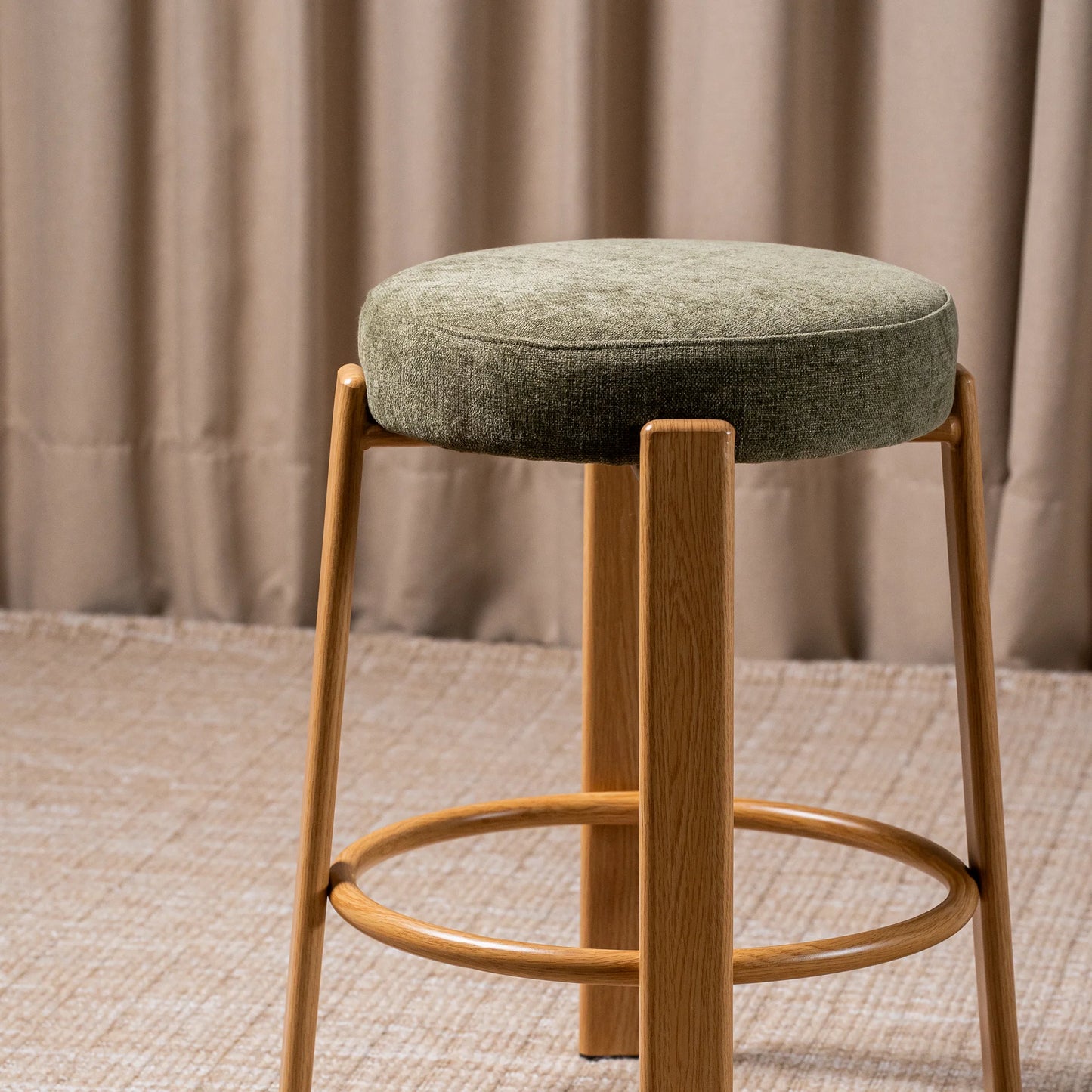 A modern bar stool with a round, textured sage green upholstered seat and a light natural wood frame with a footrest, set against a neutral beige curtain background.