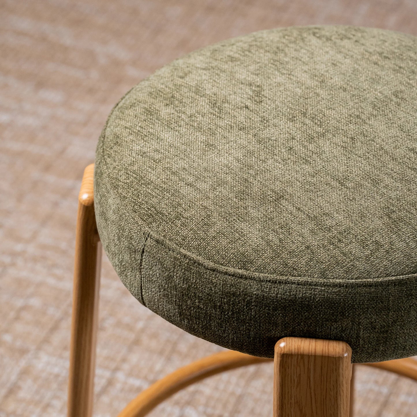 A close-up of a round upholstered stool seat in textured olive green fabric, with light natural wood legs