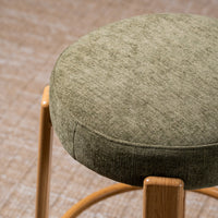 A close-up of a round upholstered stool seat in textured olive green fabric, with light natural wood legs