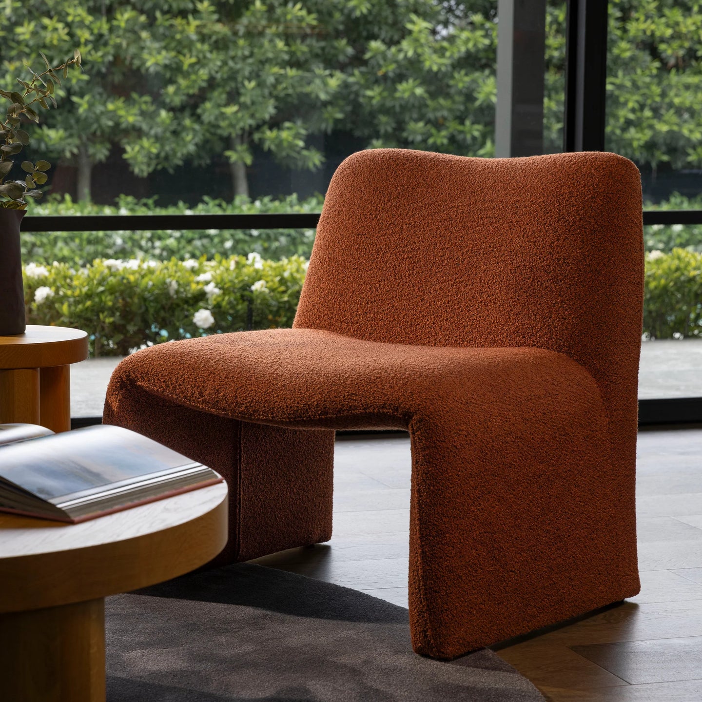 A modern sculptural armchair upholstered in a textured terracotta-brown bouclé fabric, positioned in a contemporary indoor setting with a large window showing green foliage outside.