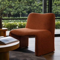 A modern sculptural armchair upholstered in a textured terracotta-brown bouclé fabric, positioned in a contemporary indoor setting with a large window showing green foliage outside.