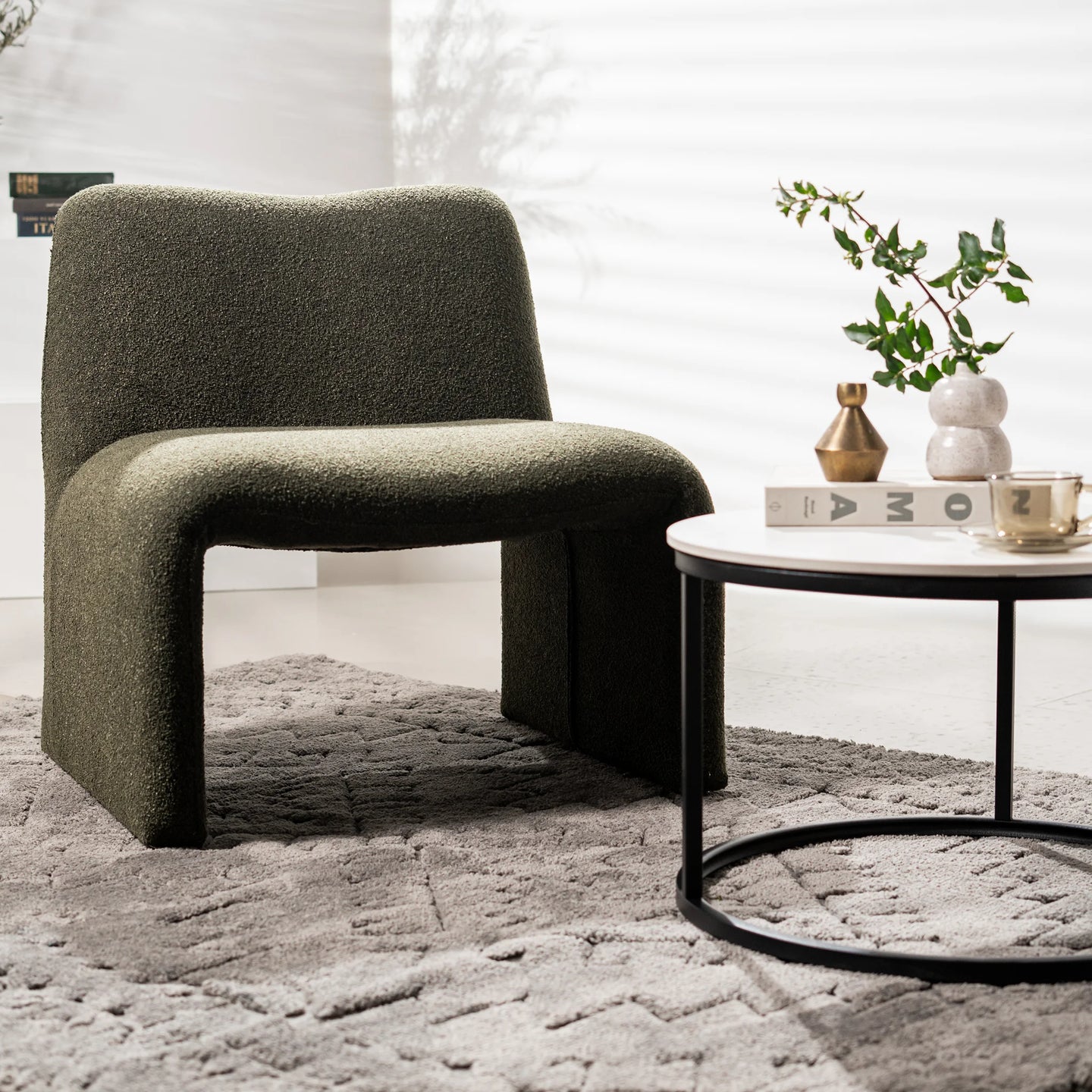 A modern upholstered lounge chair in textured olive green fabric, styled in a bright room next to a round white coffee table with a black metal frame.