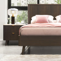 A modern bedroom scene featuring a dark brown wooden bed frame with a slatted headboard, dressed in pink bedding and pillows, next to a matching dark brown nightstand with a ribbed front and gold pull handles.