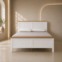 A white wooden bed frame with natural oak trim, dressed with a light beige textured duvet and two white pillows, set in a minimalist, warm-toned bedroom interior.