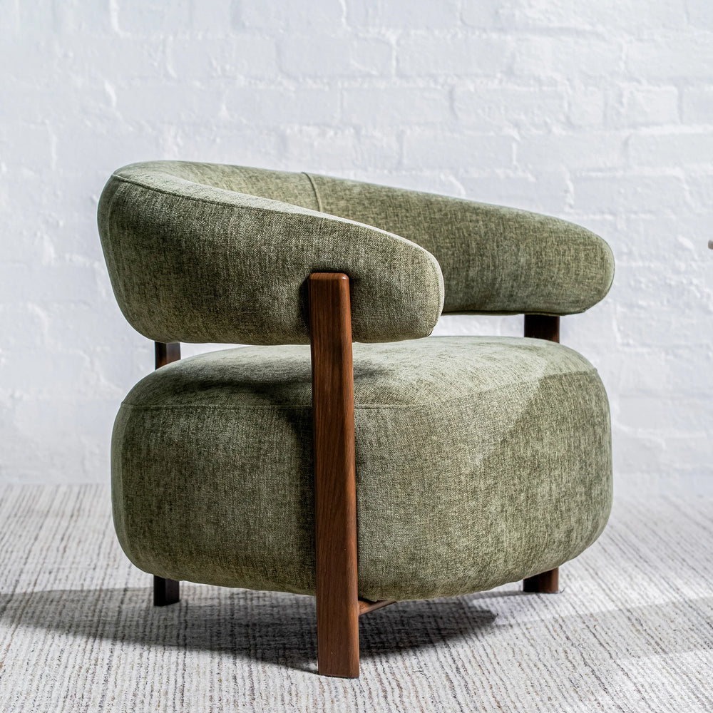 A modern barrel-style armchair upholstered in textured sage green fabric with a curved backrest, integrated armrests, and dark wood legs, set against a white brick wall and light gray rug.