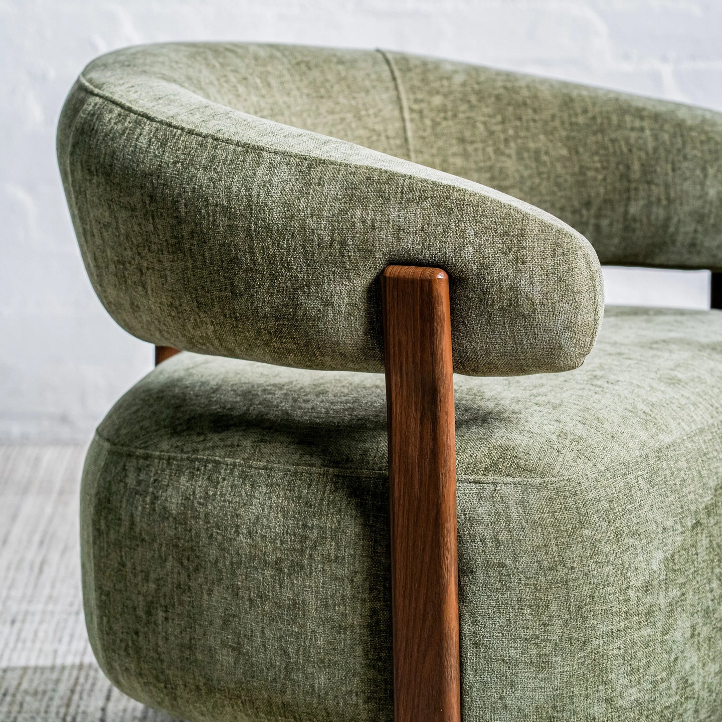 Close-up of a modern upholstered armchair with a curved light sage green fabric seat and back, supported by a warm medium-brown wooden frame leg, set against a light gray brick wall.