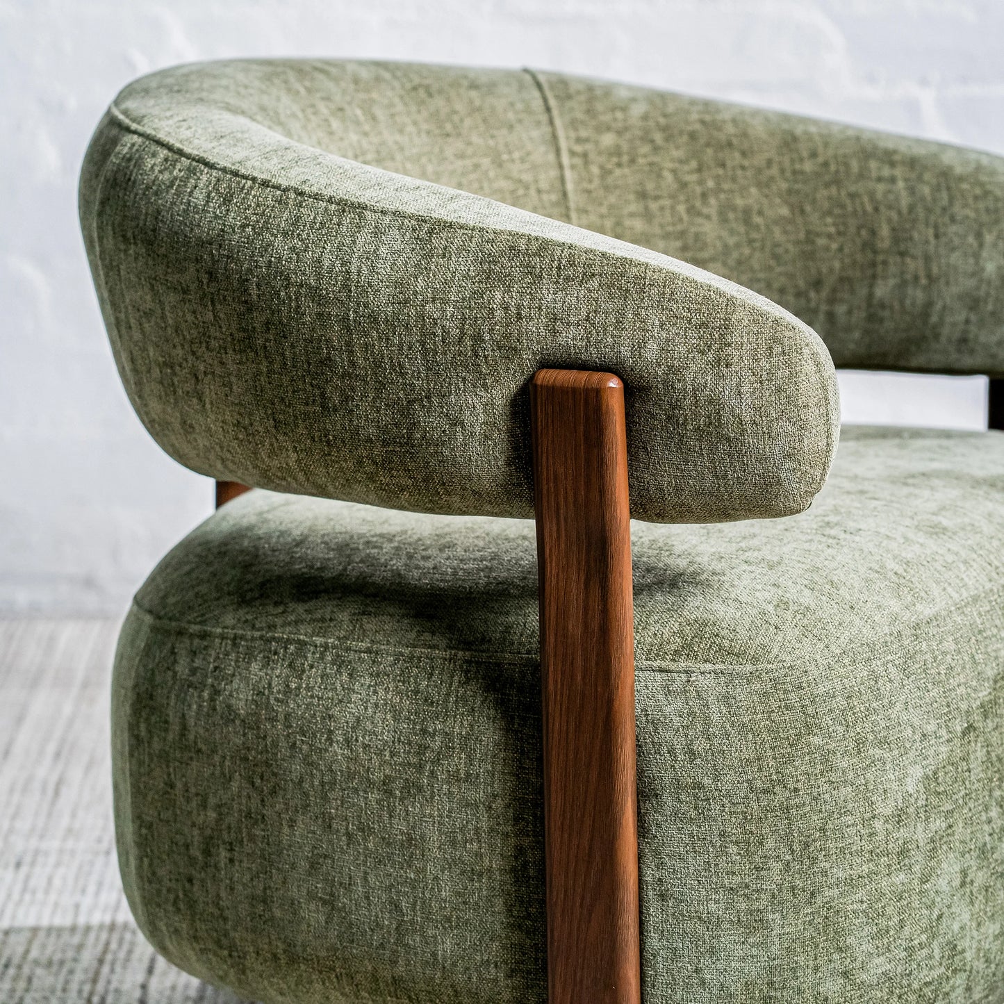 Close-up of a modern upholstered armchair with a curved light sage green fabric seat and back, supported by a warm medium-brown wooden frame leg, set against a light gray brick wall.