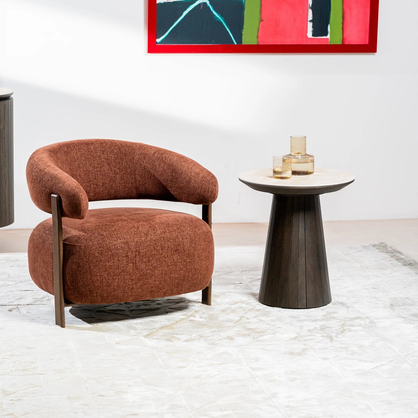 A modern curved armchair upholstered in rust-brown textured fabric with dark wood legs, positioned next to a small round side table with a light stone top and dark fluted pedestal base, set on a light marble floor against a white wall with abstract wall art.