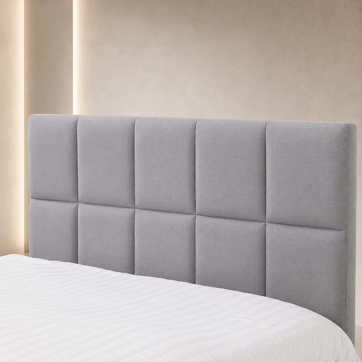A light gray upholstered headboard with a tufted grid design, shown on a bed with white bedding against a warm beige wall with soft vertical lighting.