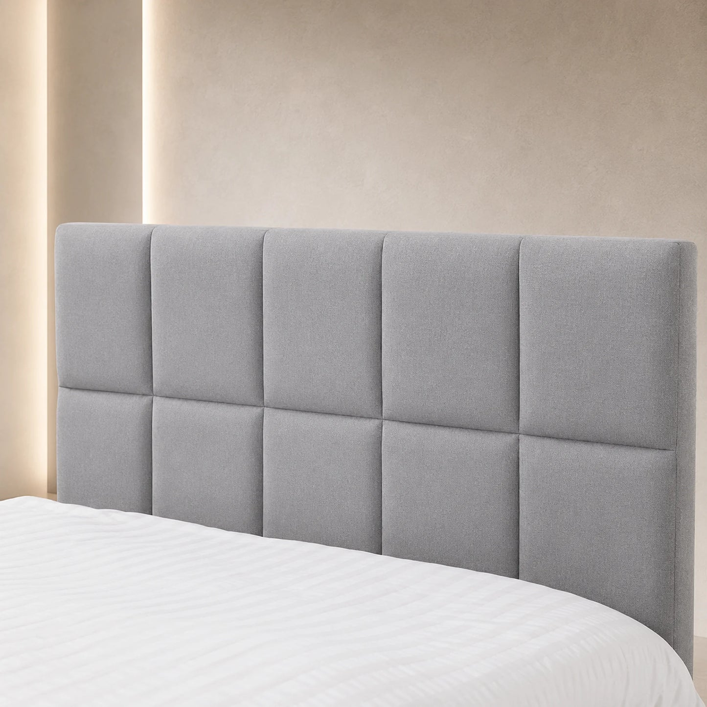 A light gray upholstered headboard with a tufted grid design, shown on a bed with white bedding against a warm beige wall with soft vertical lighting.