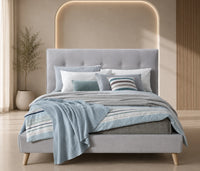 A modern light gray upholstered bed with a tufted headboard and wooden legs, styled with blue and white striped bedding, a matching blue throw blanket, and decorative pillows in a warm beige-toned bedroom.