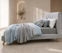 A modern light gray upholstered bed with a tall tufted headboard and tapered wooden legs, styled with blue and striped pillows and a matching blue throw blanket in a softly lit bedroom.