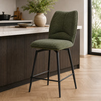 An upholstered bar stool with a textured olive green seat and backrest, supported by slim black metal legs, positioned in a modern kitchen setting.