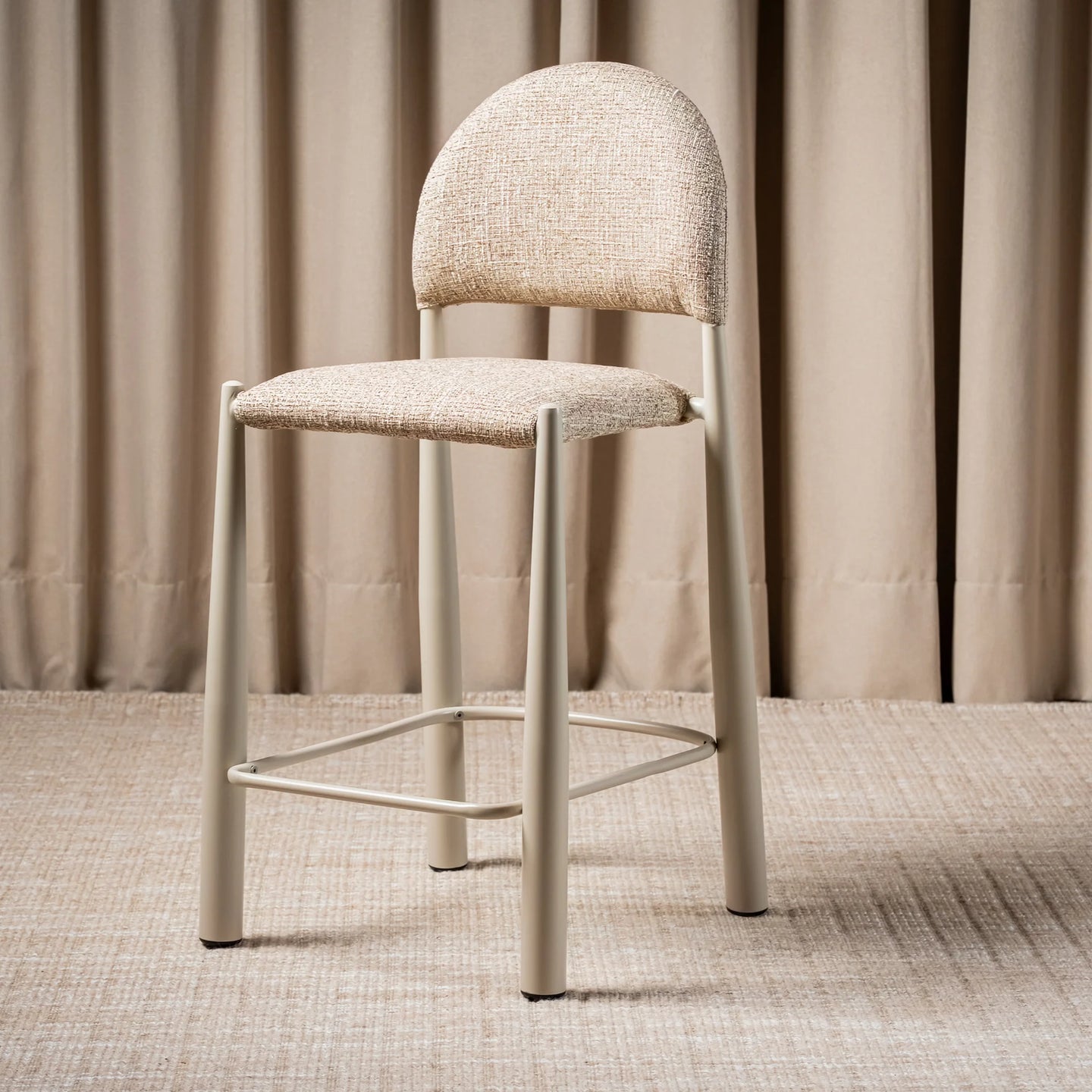 A modern bar stool with a light beige textured fabric seat and curved backrest, supported by a slim light gray metal frame with a footrest
