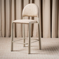 A modern bar stool with a light beige textured fabric seat and curved backrest, supported by a slim light gray metal frame with a footrest