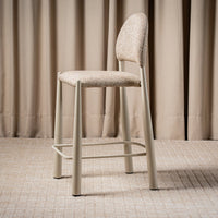 A modern bar stool with a textured beige upholstered seat and curved backrest, supported by slim light gray metal legs and a footrest