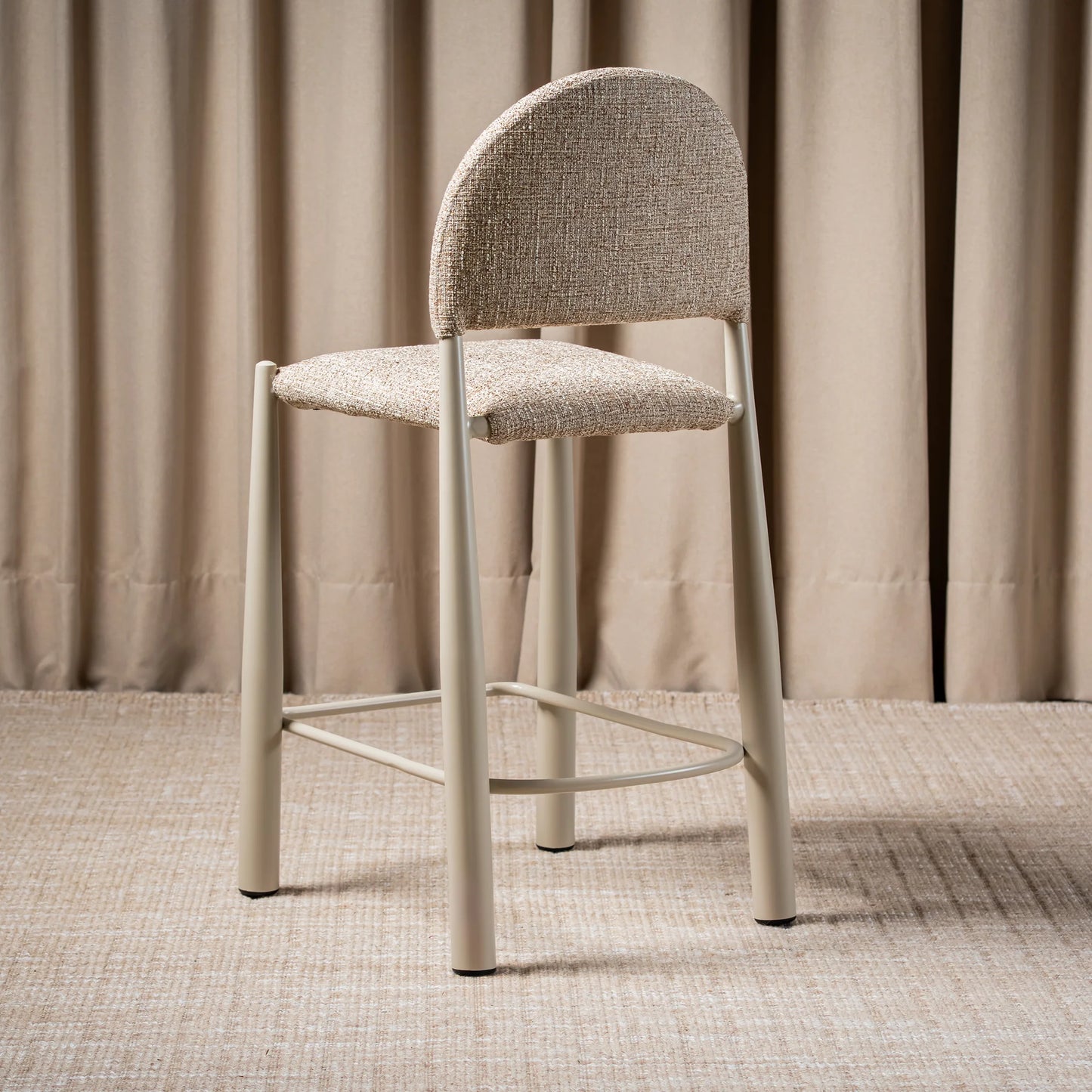 A modern counter stool with a textured beige upholstered seat and curved backrest, supported by slim light gray metal legs