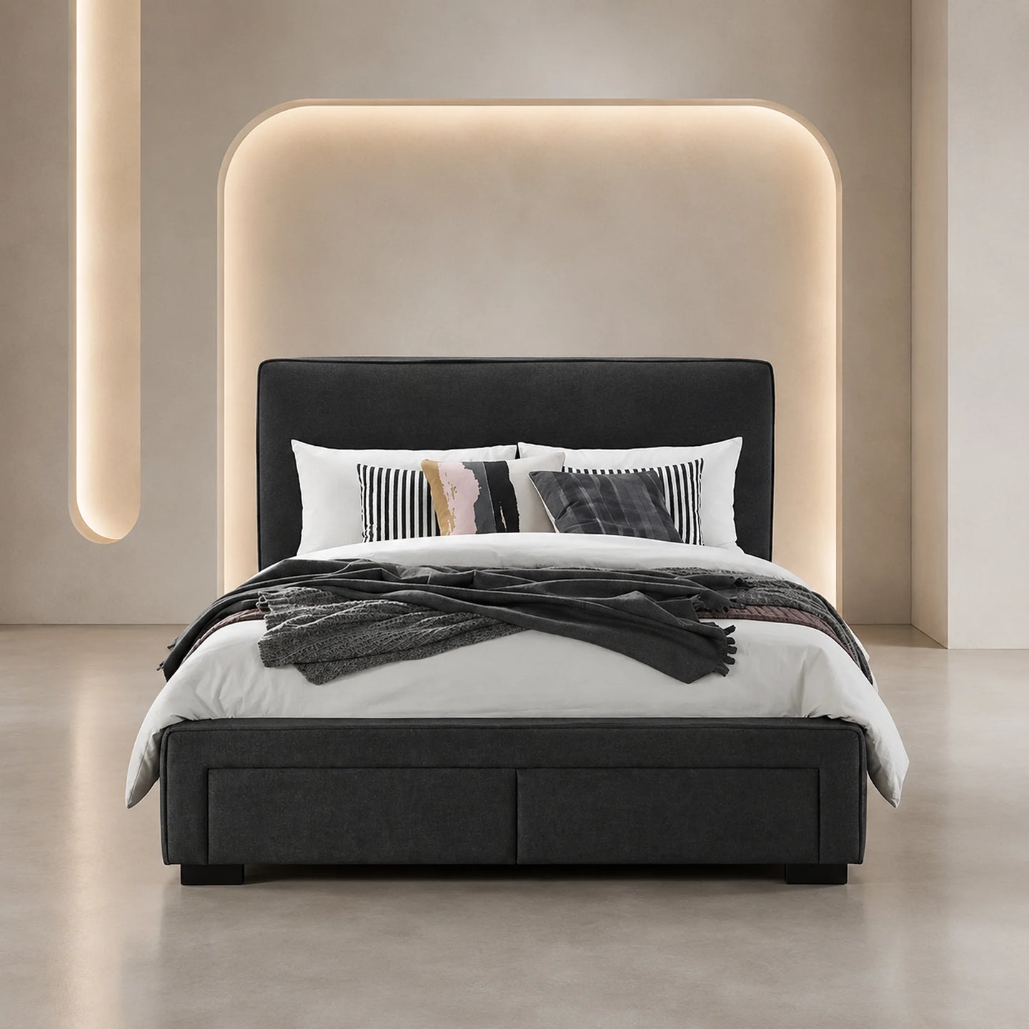 A modern platform bed with a dark charcoal upholstered headboard and footboard, styled with white bedding, striped and patterned accent pillows, and a dark throw blanket, set against a minimalist beige wall with integrated warm lighting.
