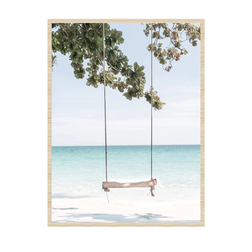 A framed art print depicting a wooden swing hanging from a tree over a tropical beach with turquoise water and white sand, set against a light gray background.