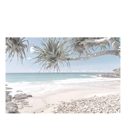 A coastal beach scene with pale sand, pebbles, turquoise water, and a pale blue sky, framed by overhanging leafy branches in the foreground.